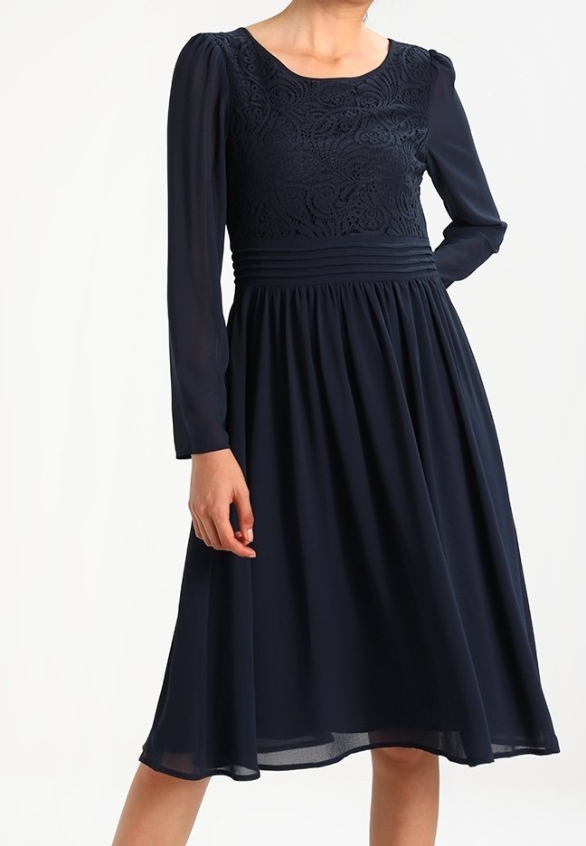 Femme portant une robe midi bleu marine à manches longues, corsage en dentelle, taille plissée et jupe évasée, debout avec une main sur la hanche.