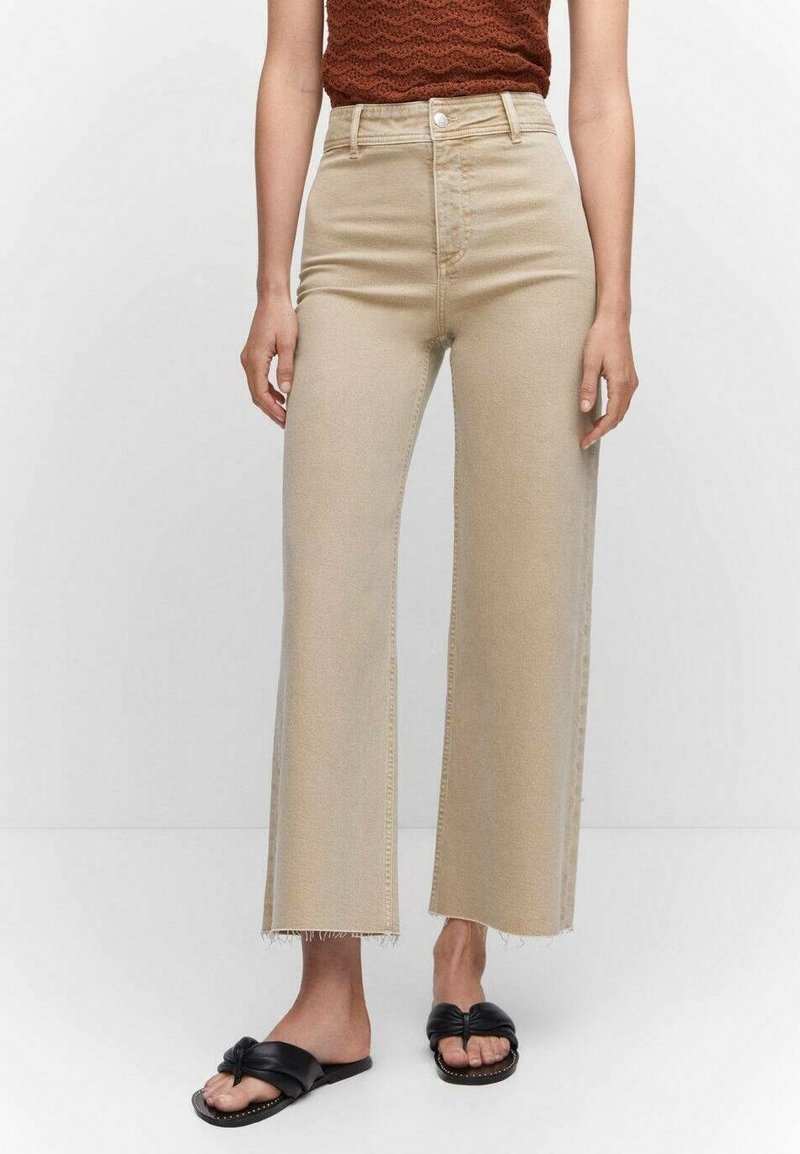 Femme portant un jean beige taille haute à jambes larges, associé à des sandales plates noires avec nœud et un haut marron texturé sans manches.