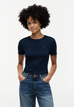 Γυναίκα με σγουρά μαλλιά, φορώντας μπλε κοντομάνικο εφαρμοστό μπλουζάκι και ψηλόμεσα μπλε τζιν, με τα χέρια στις τσέπες.