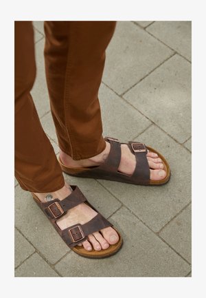Brune lær sandaler med to justerbare stropper og metalspænder, hvilende på en stenbelagt overflade, parret med brune bukser.