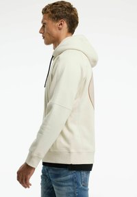 Crème-kleurige hoodie met een gestructureerde pasvorm, capuchon en contrasterende zwarte trekkoorden. Beschikt over een textuurdetail op de achterkant.