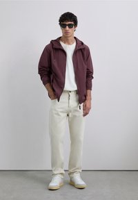 Mand i sorte solbriller, maroon zip-up jakke, hvid skjorte, beige bukser og hvide sneakers står mod en ensfarvet grå væg.