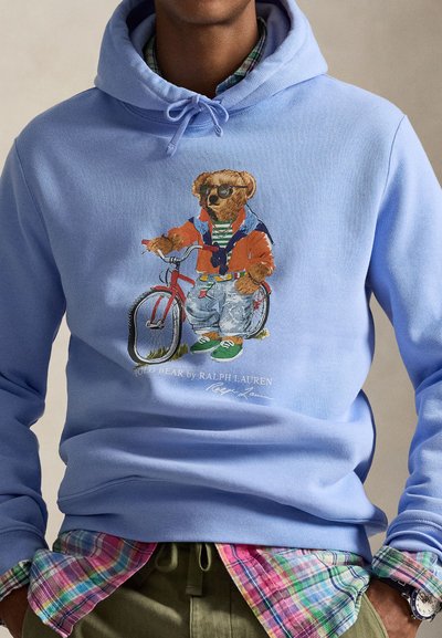 Ανοιχτό μπλε φούτερ με κουκούλα που απεικονίζει μια αρκούδα να οδηγεί ένα κόκκινο ποδήλατο, φορώντας πορτοκαλί μπουφάν και τζιν. Κάτω από αυτό φαίνεται πουκάμισο με καρό μοτίβο, σχέδιο casual.