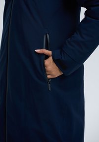 Marine buitenkleding met een gladde textuur, voorzien van een ritszak en klittenbandaccenten bij de manchetten. Eenvoudig ontwerp met functionele details.