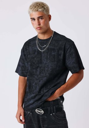 Jonge man met kort blond haar draagt een zwart ""Change The Game"" T-shirt, gelaagde kettingen, zwarte jeans en een riem met een zilverkleurige gesp.