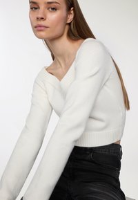 Maglione bianco a maniche corte con scollatura a V e polsini a costine, realizzato in tessuto morbido. Indossato con pantaloni neri a vita alta, mettondo in risalto il contrasto dell'outfit.
