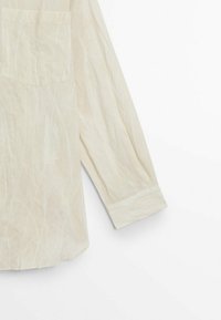 Chemise beige clair à manches longues avec des plis visibles, une poche poitrine et des poignets boutonnés sur fond blanc.