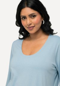 Hellblaues Oberteil mit Rundhalsausschnitt und langen Ärmeln, hergestellt aus weichem Stoff; zeichnet sich durch eine glatte Textur und eine lockere Passform für hohen Tragekomfort aus.