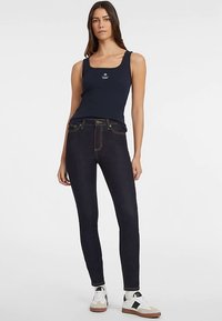 Marineblauw tanktop met logo, gecombineerd met donkerblauwe high-waisted jeans van denim met gele stiksels en witte sneakers met zwarte accenten.