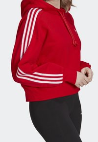 Red cropped hoodie in cotton blend with white three-stripe design on sleeves, adjustable drawstring, and a relaxed fit.