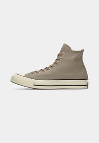 Adidași înalți din piele sintetică bej, cu vârf din cauciuc, talpă intermediară albă în contrast și șireturi maronii, având branding clasic pe călcâi.