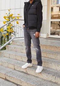 Svart pufferväst över en svart långärmad tröja, grå slitna jeans och vita sneakers. Står på stentrappor med gula blommor.