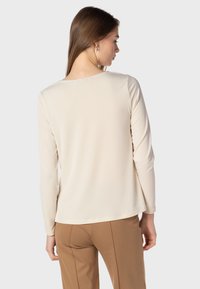 Langärmeliges cremefarbenes Oberteil mit rundem Ausschnitt und glatter Textur. Kombiniert mit braunen Hosen, die eine figurnahe Silhouette betonen.