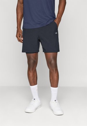 Shorts de sport marine en tissu léger, avec un devant plat et des poches latérales. Portés avec des baskets blanches et des chaussettes mi-mollets.