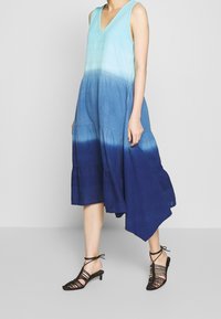 Robe sans manches en V avec un dégradé du bleu clair au bleu foncé. Présente un tissu texturé et une coupe fluide avec un ourlet en bas en étages. Sandales noires à brides.
