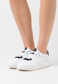 Vita atletiska sneakers med svarta accenter, perforerad textur, vadderad tunga, tjock gummisula och märkesetikett på sidan.