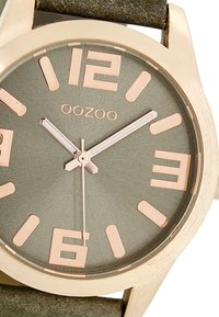 Montre métallique ronde avec un boîtier doré, un cadran gris, de grands index en or rose et un bracelet en tissu marron. Logo présent en haut.