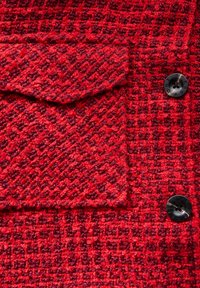 Red tweed fabric with a houndstooth pattern, featuring a patch pocket and two black buttons on the front. Textured, woven design.