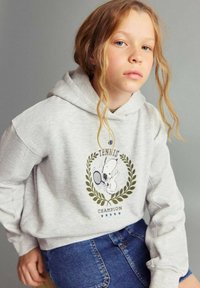 Mladé dievča s svetlými kučerami, oblečené v šedej mikine s potlačou Snoopyho tenisového šampióna a modrej denimovej sukni, sediace na jednoduchom pozadí.