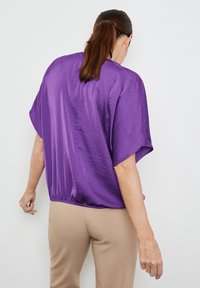 Lila satin topp med korta, breda ärmar och en rynkad nederdel. Bärs med beige byxor. Slät textur och glänsande yta.