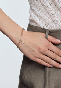 Złota bransoletka łańcuszkowa z zawieszką w kształcie kwiatu, składająca się z połączonych owalnych elementów o gładkiej fakturze. Noszona na ręce na neutralnym tle.