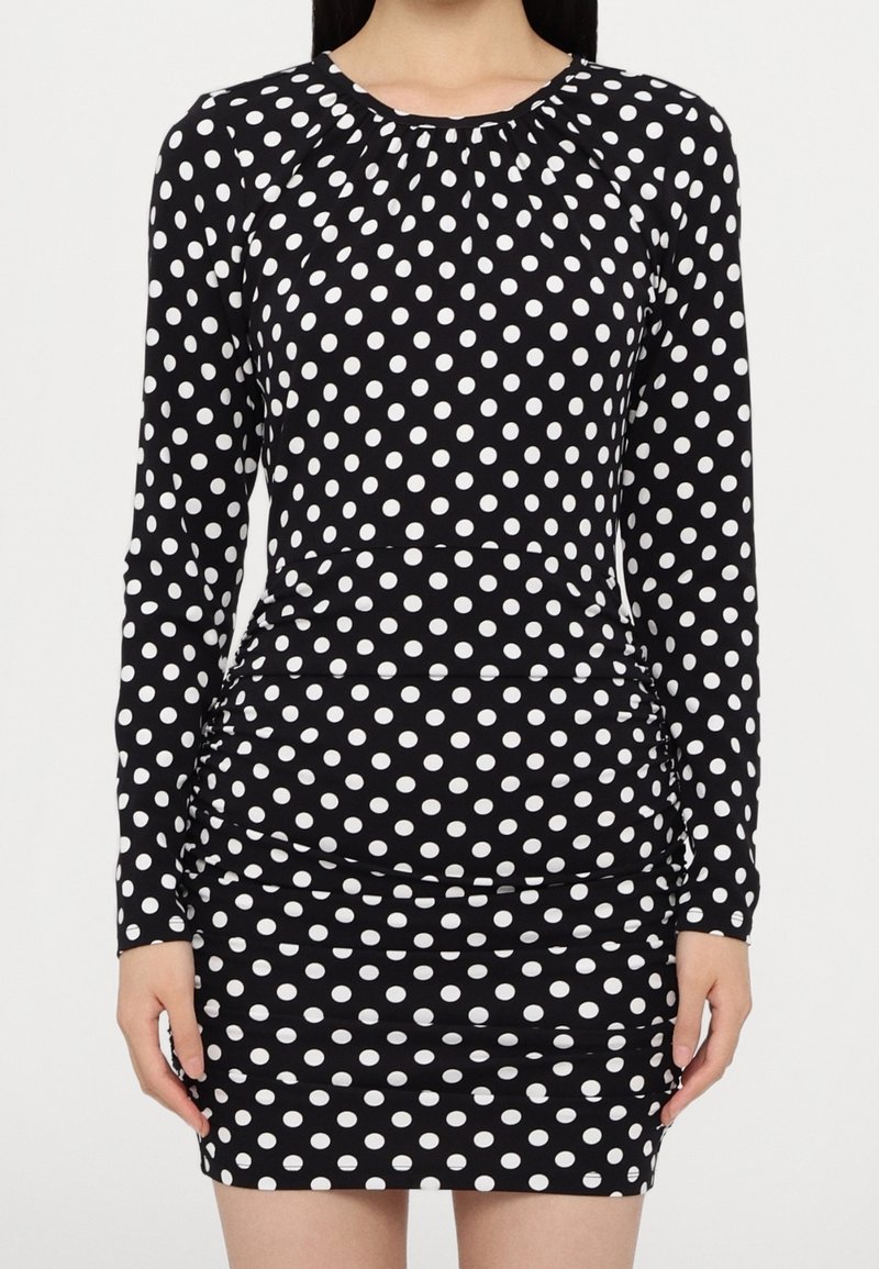 Femme portant une robe noire cintrée à manches longues avec des pois blancs, debout, les bras détendus le long du corps.