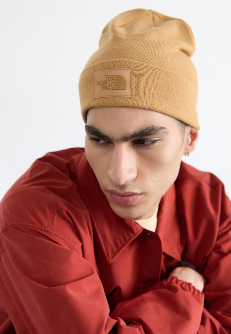 Beige gestrickte Mütze mit einem rechteckigen Lederpatch, das das eingeprägte Logo "THE NORTH FACE" zeigt. Getragen von einer Person in einer roten Jacke.