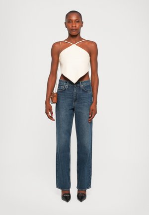Crème asymmetrische top met kruislings bandjes, gecombineerd met blauwe wijde jeans. Model draagt zwarte puntige hakken en heeft een kleine bruine tas.