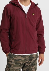 Bordowa lekka kurtka z kapturem, zapinana na zamek, z kieszeniami bocznymi. Posiada elastyczne mankiety i małe żółte logo na klatce piersiowej.