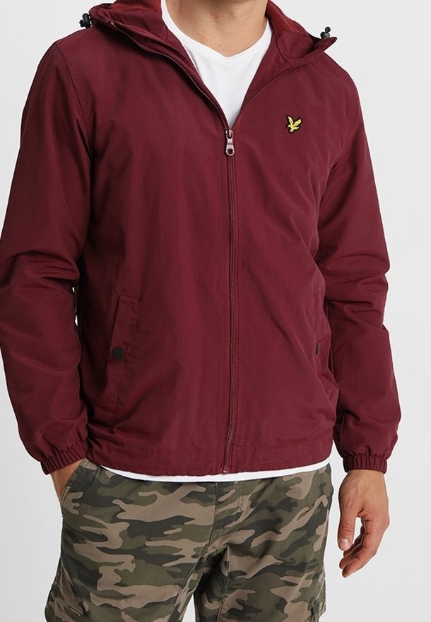 Bordowa lekka kurtka z kapturem, zapinana na zamek, z kieszeniami bocznymi. Posiada elastyczne mankiety i małe żółte logo na klatce piersiowej.