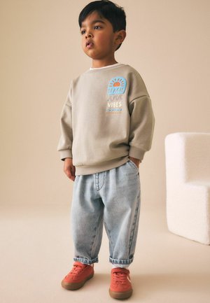 Young child wearing beige sweatshirt with "Beach Vibes" print, light blue rolled-up jeans, and orange sneakers, standing with hands in pockets.
