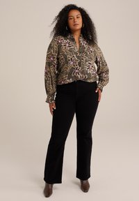 Blusa floreale verde con colletto, maniche lunghe e motivi intricati. Abbinata a pantaloni svasati neri e scarpe marroni a punta.