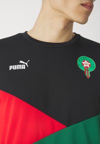 Camiseta deportiva negra con acentos diagonales en rojo y verde. Presenta un logo de Puma en blanco y un colorido emblema marroquí. Tejido texturizado.