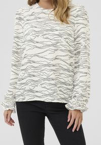 Biała bluzka z długim rękawem z czarnym falistym wzorem, okrągłym dekoltem i marszczonymi mankietami, nadająca fakturowany wygląd. Połączona z czarnymi spodniami.