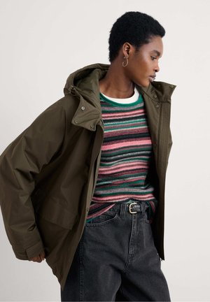 Olivegrüne Jacke mit Kapuze und Druckknöpfen über einem mehrfarbig gestreiften Pullover. Das Model trägt schwarze hochgeschnittene Jeans mit einem geflochtenen Gürtel.