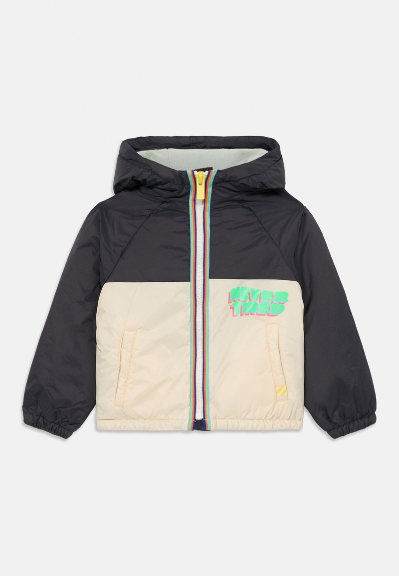 Polsterdatud jakk, mille ülaosa on must ja alumine osa ko cream, varustatud mitmevärvilise tõmblukuga ning "NEVER TIRED" graafikaga esiküljel.