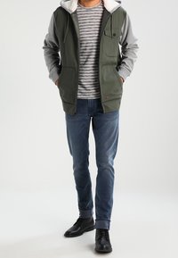 Gilet verde con maniche grigie, cappuccio foderato, tasche frontali e dettaglio con bottoni, indossato sopra una camicia a righe grigie e jeans blu. Scarpe nere.