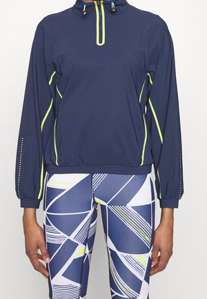 Granatowa sportowa bluzka z długim rękawem z neonowożółtym obszyciem, w komplecie z legginsami w geometryczny wzór w kolorach granatowym, białym i neonowożółtym.