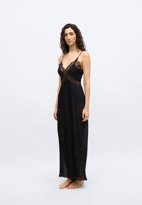 Γυναίκα με μακριά σγουρά μαλλιά, φορώντας ένα αμάνικο, μαύρο δαντελένιο και πιετέ φορεματάκι μέχρι τον αστράγαλο, όρθια ξυπόλητη πάνω σε ένα απλό φωτεινό φόντο.