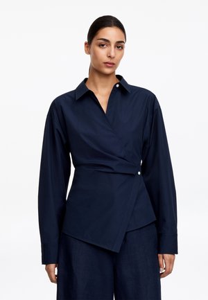 Femme aux cheveux foncés attachés, portant un chemisier bleu marine à manches longues style portefeuille et un pantalon large assorti sur fond blanc.