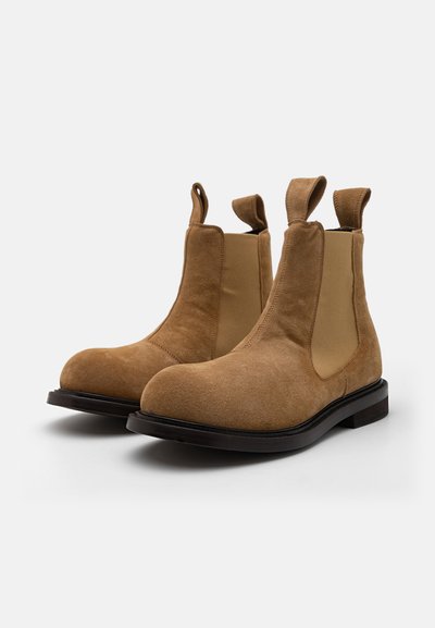Botas Chelsea de ante en tono marrón claro, con paneles laterales elásticos, punta redondeada, tiradores y suela plana negra. Parte superior texturizada con acentos lisos.