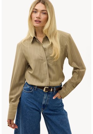 Femme blonde portant une chemise beige à fines rayures boutonnée, rentrée dans un jean bleu taille haute avec une ceinture foncée, posant avec une main dans la poche.
