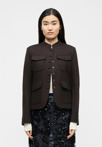 Γυναίκα με μακριά σκούρα μαλλιά που φοράει σκούρο καφέ σακάκι με κουμπιά και τέσσερις τσέπες με καπάκια πάνω από λευκή μπλούζα και μαύρη φούστα με παγιέτες.