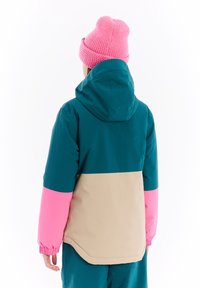 Sak s blokovými barvami s modrozelenou horní částí, béžovým středem a růžovými dolními rukávy. Funkce kapuce a elastické manžety. Na hlavě pletená růžová čepice.