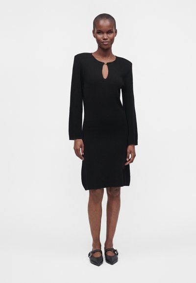 Femme portant une robe noire longueur genoux avec manches longues, un décolleté en trou de serrure à l'avant, et des chaussures Mary Jane noires à bout pointu.