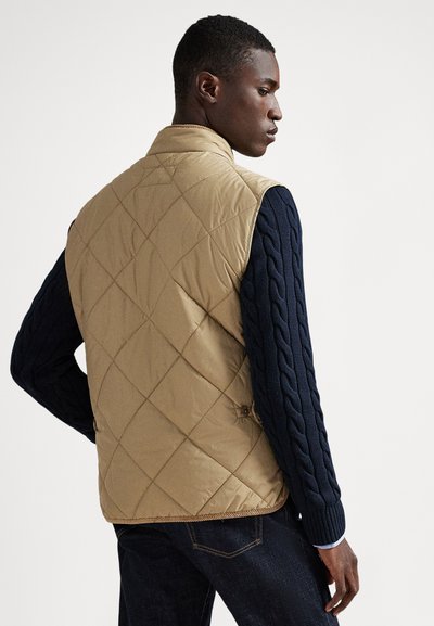 Beżowa pikowana kamizelka z wysokim kołnierzem, zestawiona z granatowym swetrem w drobny warkocz. Charakteryzuje się detalami na guzikach i fakturowanym wykończeniem.