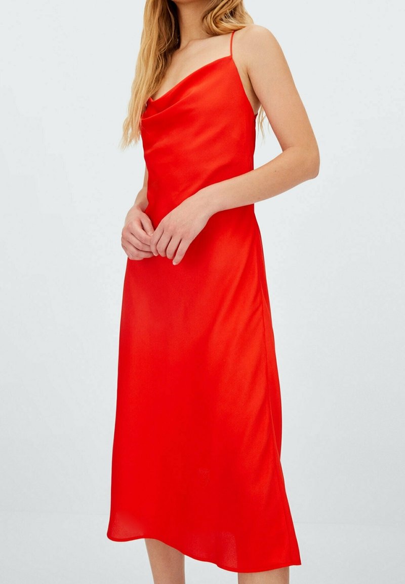 Femme portant une robe midi rouge vif sans manches avec fines bretelles et un décolleté drapé, debout les mains légèrement jointes.