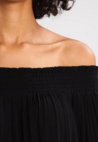 Top nero a spalle scoperte con scollatura elastica smock e tessuto fluido e testurizzato. Caratterizzato da un design leggero e arricciato.