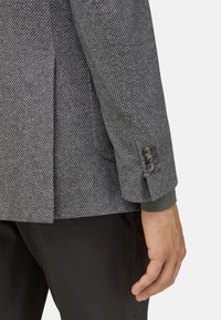 Texturovaný šedý blazer s jemným vzorem herringbone, s jedním knoflíkem a kontrastními pletenými manžetami. Černé kalhoty viditelné dole.
