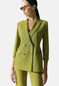 Giacca doppiopetto in tessuto verde brillante, con revers a punta, maniche arrotolate e pantaloni su misura abbinati.